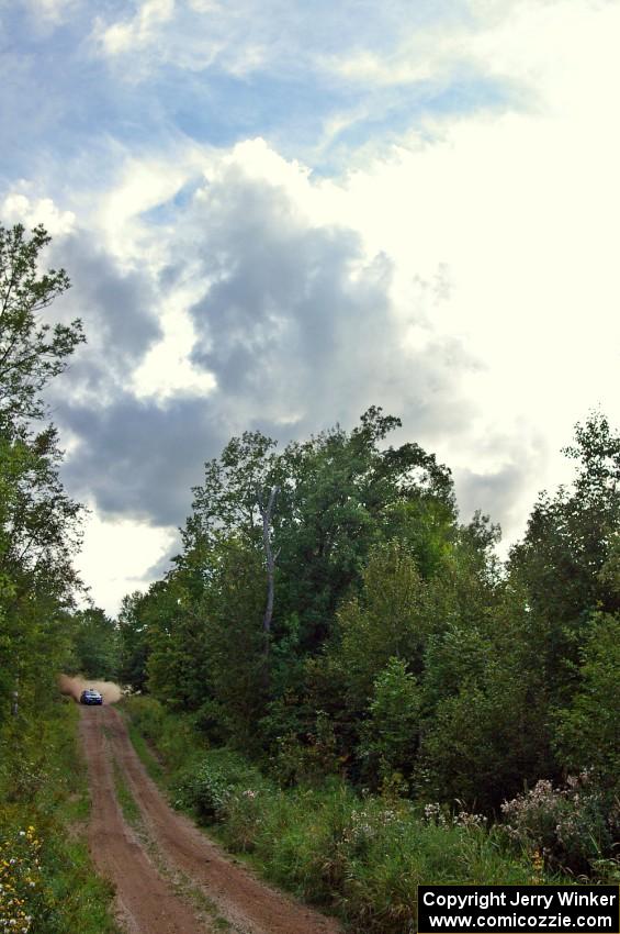 The Ken Block / Alex Gelsomino Subaru WRX STi rockets down a straight under gorgeous cloud-covered skies.